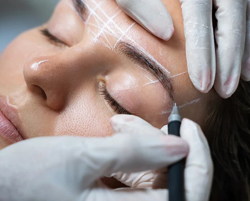 Close-up of a woman undergoing a microblading procedure, with white pre-drawing lines on her eyebrows and a technician using a pen-like tool to enhance the shape.